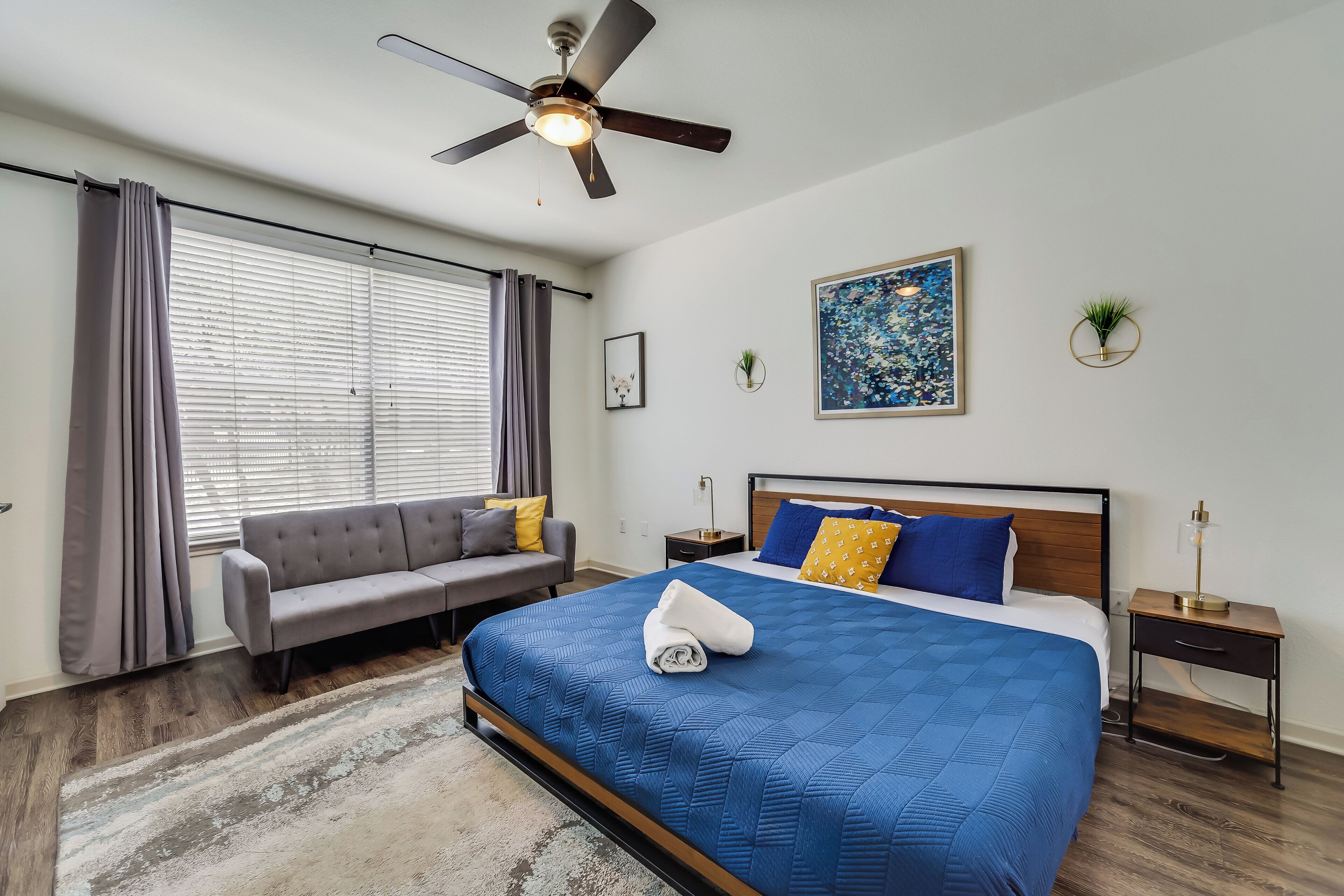 A bedroom with a bed, a sofa, a ceiling fan, a window with curtains, a painting, and a small plant.