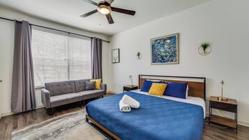 A bedroom with a bed, a sofa, a ceiling fan, a window with curtains, a painting, and a small plant.