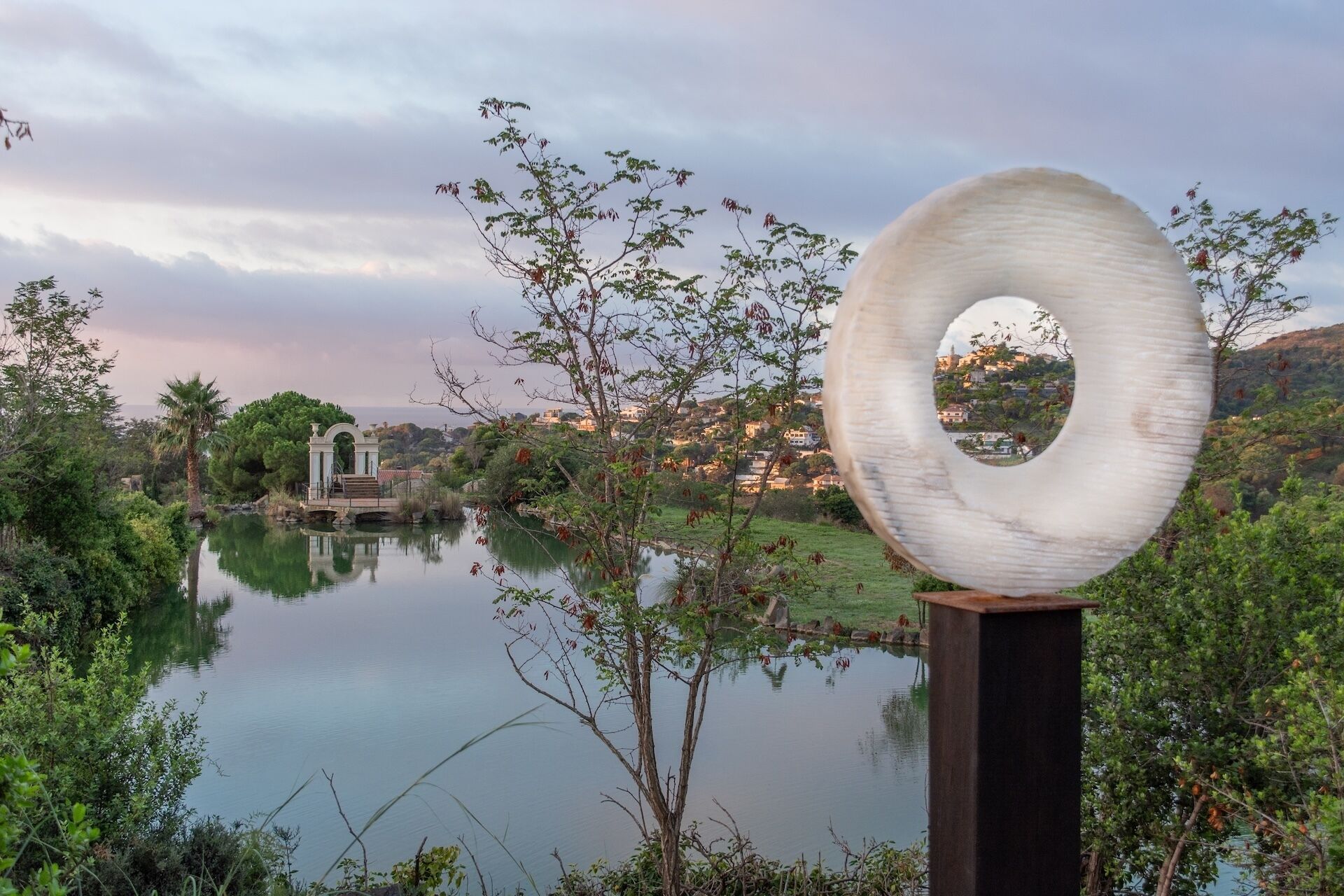 A serene lakeside view with a unique circular sculpture in the foreground, a bridge, and a distant town.