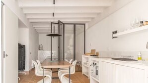 A modern kitchen with white cabinetry, a dining area with a wooden table and white chairs, and a large window providing natural light. - Romantic apartment in idyllic nature setting (Barcelona)