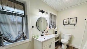 Une salle de bain avec un meuble-lavabo blanc, un miroir rond, des toilettes et une fenêtre avec des rideaux.