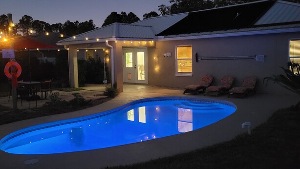 A swimming pool with a blue tint, surrounded by a patio area with chairs and a small table. - Your Palm Coast Pool House! (Palm Coast)