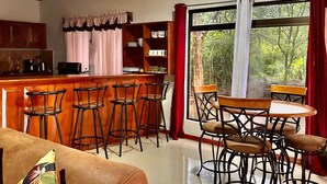 Ein gemütlicher Wohnbereich mit einer Couch, einem Couchtisch und einem Esstisch mit Stühlen. Eine Küchenzeile mit Bar und Hockern ist ebenfalls vorhanden. Große Fenster mit Vorhängen bieten Blick auf Grünflächen.