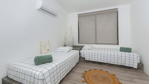 Una habitación pequeña y moderna con dos camas, una ventana con persiana, un tapete pequeño y un equipo de aire acondicionado.
