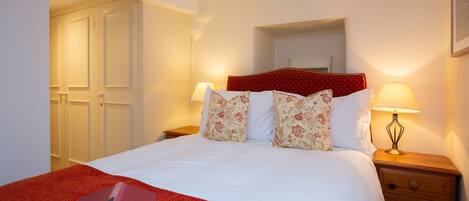 A neatly made bed with floral pillows, a red throw blanket, and a book on the bed. There is a nightstand with a lamp and a wooden drawer. A door and a mirror are visible in the background.