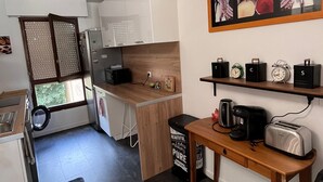 A compact kitchen with white cabinets, a wooden countertop, and a black trash bin. There is a window with a view of greenery outside, and a shelf with clocks and a framed picture above the counter. - Close to Dijon city center, ideal for a leisure or business stay  (Dijon)