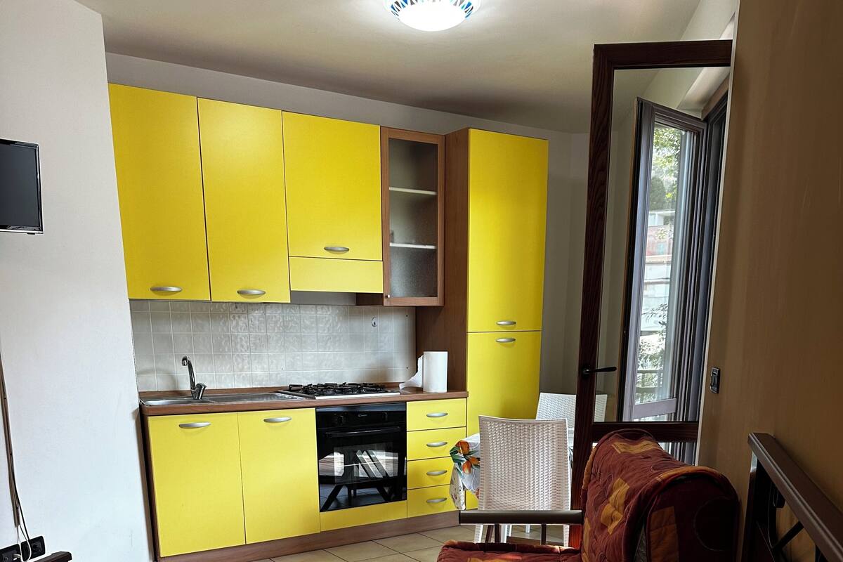 Una habitación compacta con cama, un pequeño sofá, una mesa de comedor y una kitchenette con armarios amarillos.