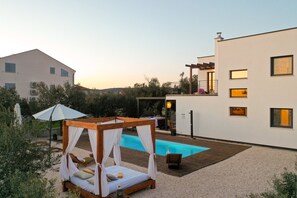 A modern outdoor pool area with a wooden deck, a canopy bed, and a white building in the background. - Villa Fusion with Pool (Sukošan)