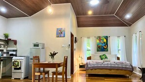 A bedroom with a bed, a dining table with chairs, a kitchenette with a microwave and oven, and a painting on the wall.