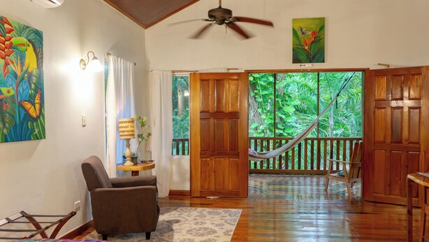 A spacious room with a wooden floor, a hammock, a chair, and a door leading to a balcony with a view of greenery.