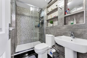 Un baño moderno con ducha envuelta en vidrio, inodoro blanco, lavamanos y repisas de mármol con artículos de aseo.