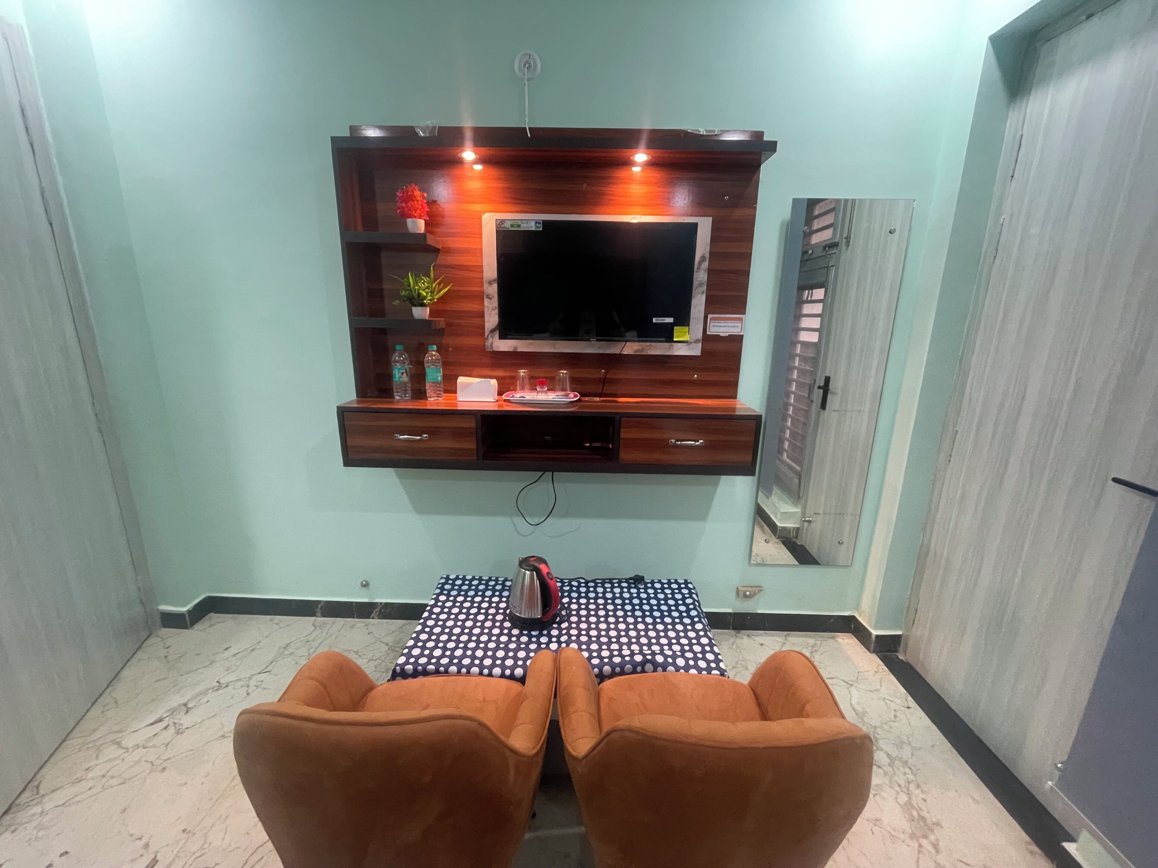 A room with a TV mounted on a wooden wall, a small table with a kettle, two orange chairs, and a mirror on the wall.