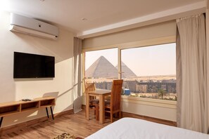 A hotel room with a bed, a desk, a chair, and a large window offering a view of the pyramids. - Rosetta pyramids inn (Giza)