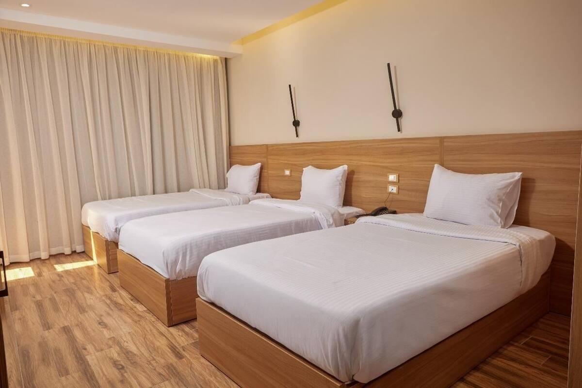 Habitación de hotel con dos camas individuales, cabeceros de madera, un ventanal con cortinas y un reloj de pared.