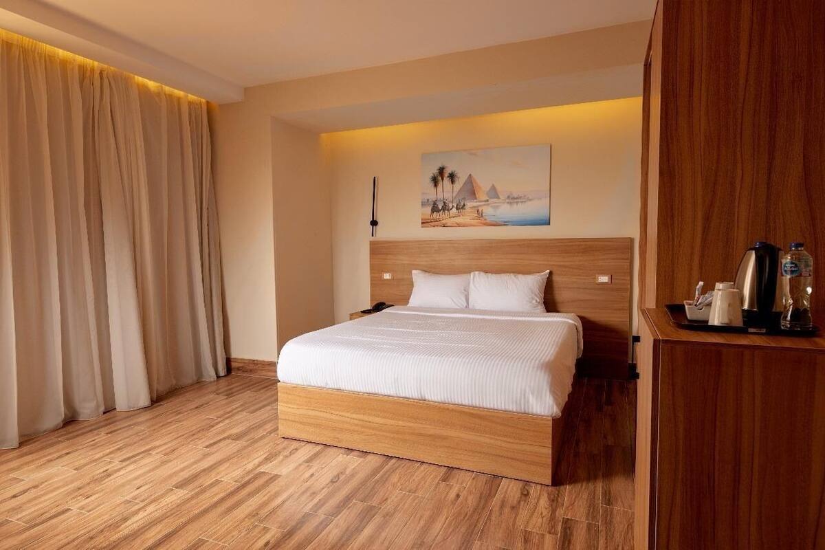 Habitación de hotel con una cama de madera, un cuadro de pirámides, una mesita de noche y un escritorio de madera con una cafetera.