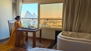 A hotel room with a large window, a dining table with food, a bathtub, and a bed.