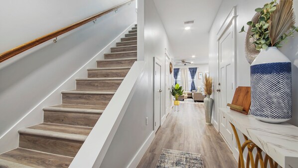 Una escalera moderna con peldaños de madera y barandilla blanca, que conduce a una sala de estar con sofá, mesa de centro y plantas decorativas.