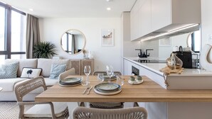 A modern living room with a wooden dining table set for four, a sofa with cushions, a round mirror, and a kitchen area with a stove and cabinets.