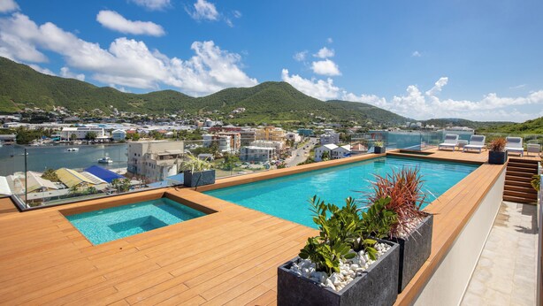 Piscina en la azotea con cubierta de madera, rodeada de plantas en macetas, con vistas a un pueblo costero y colinas.