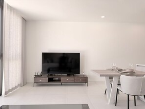 A modern living room with a flat-screen TV on a wooden stand, a dining table with chairs, and a large window with curtains.