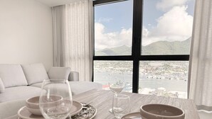 Mesa de comedor dispuesta con platos, fuentes y copas de vino, con vistas a un puerto deportivo y montañas a través de amplios ventanales.