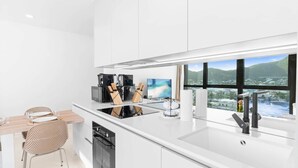 Una cocina moderna con armarios blancos, horno empotrado, microondas y cafetera. Hay un comedor con mesa y sillas de madera.