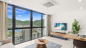 A modern living room with a large sliding glass door, a flat-screen TV, a wooden coffee table, and a view of a cityscape and mountains. - B237 - Cosy 1 Bedroom with Lagoon View (Sint Maarten)