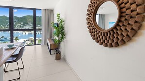 A modern room with a large window offering a view of a marina and hills. There's a wooden-framed mirror on the wall and a dining area with a table and chairs. - B237 - Cosy 1 Bedroom with Lagoon View (Sint Maarten)