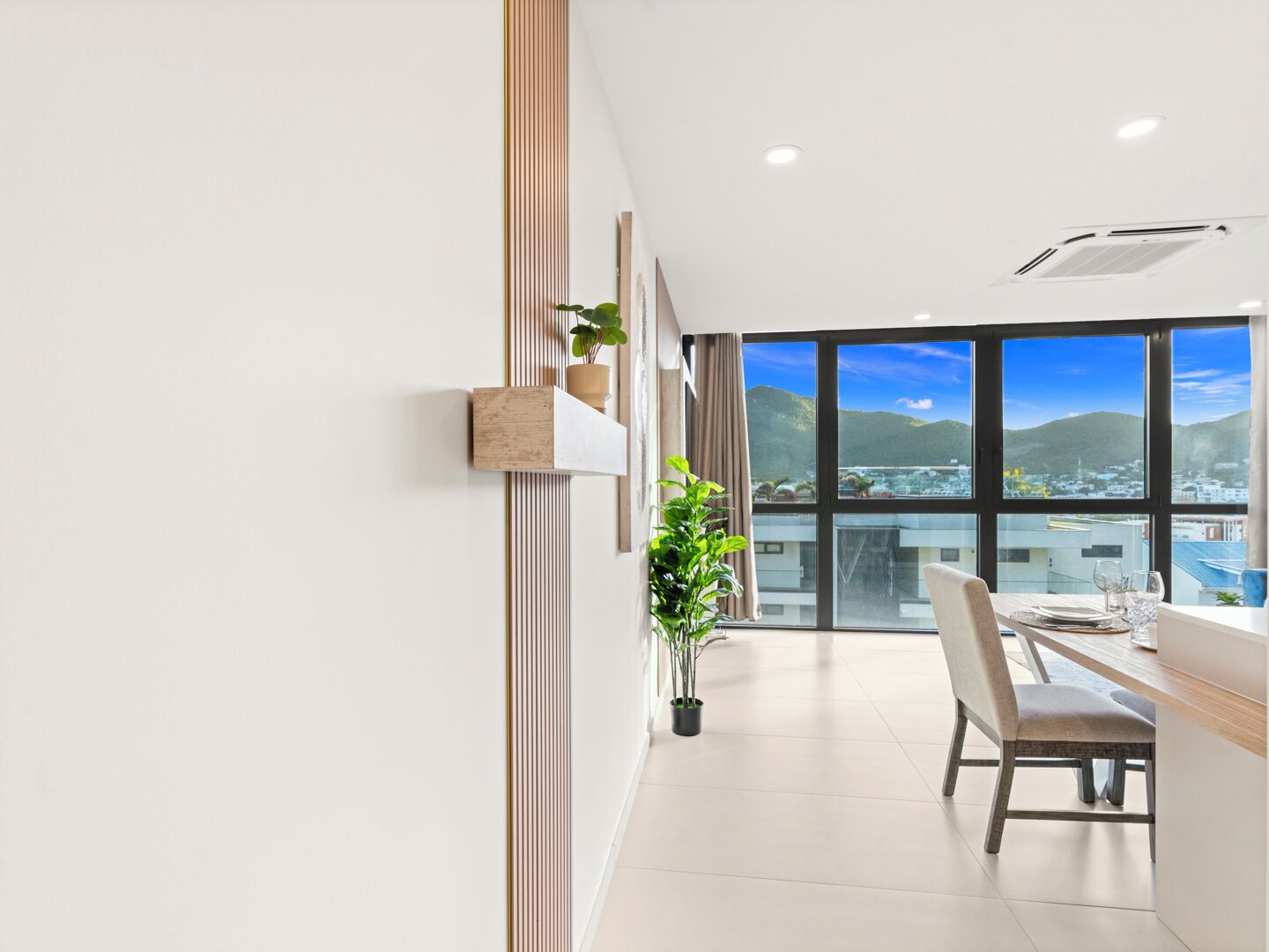 A modern hotel room with a large window offering a city view, a dining area with a table and chairs, and a potted plant.