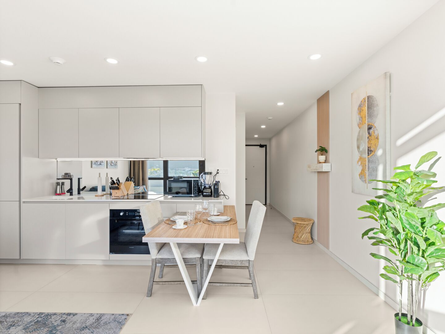A modern kitchen with white cabinets, a dining table with chairs, a microwave, and a blender. There is a plant on the right and a painting on the wall.