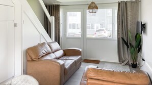 A modern living room with a brown leather sofa, a white armchair, and a small table. A large window with curtains and a view of the street is visible. - Great property close to Freshwater East Bay, perfect for a family break. (Freshwater East)
