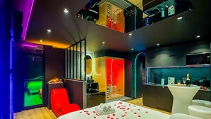 Ein modernes Hotelzimmer mit einem Bett, einem roten Sessel, einem kleinen Tisch mit Flasche und Glas sowie einer Küchenzeile.