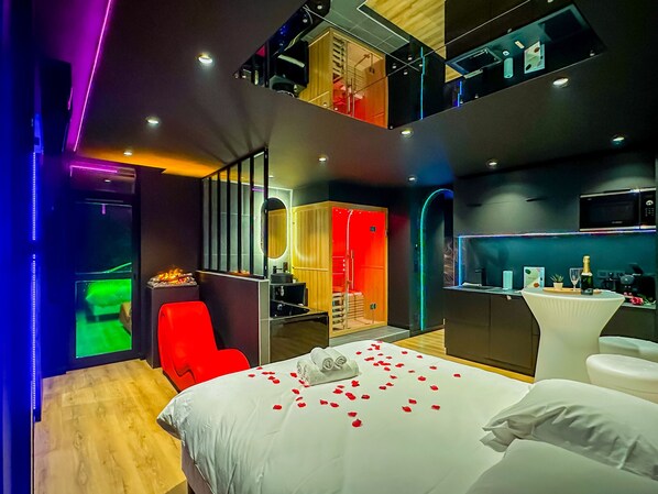 Une chambre d’hôtel moderne avec un lit, un fauteuil rouge, une petite table avec une bouteille et un verre, et un coin cuisine.