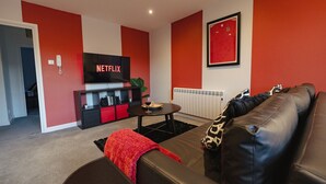 Una sala de estar con un sofá de cuero negro, una manta roja, una mesa de centro, un televisor con el logotipo de Netflix, un cuadro en la pared y un radiador.