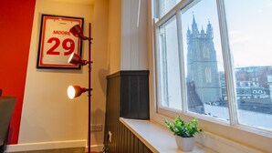 Una habitación con un cartel enmarcados con el número 29, una lámpara de pie, una planta en maceta y una ventana con vistas a un edificio histórico.
