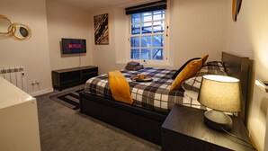 Un dormitorio con cama, mesita de noche, lámpara, televisor y ventana.