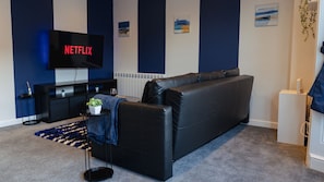 A living room with a black leather sofa, a flat-screen TV displaying 'NETFLIX,' a small side table with a plant, and a blue and white patterned rug. - Castle Views One Bed Apartment (Cardiff)