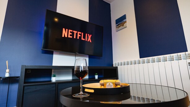 Een kamer met een televisie waarop 'NETFLIX' staat, een glas rode wijn, een dienblad met Ferrero Rocher chocolade en een blauw-wit kleurenschema.