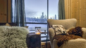 Une pièce chaleureuse avec une grande fenêtre, un fauteuil recouvert d’un plaid moelleux, une petite table avec une bougie, et une vue sur un paysage enneigé.