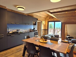 Une cuisine moderne avec des meubles de couleur sombre, une table à manger en bois et un coin salon confortable offrant une vue sur les montagnes.