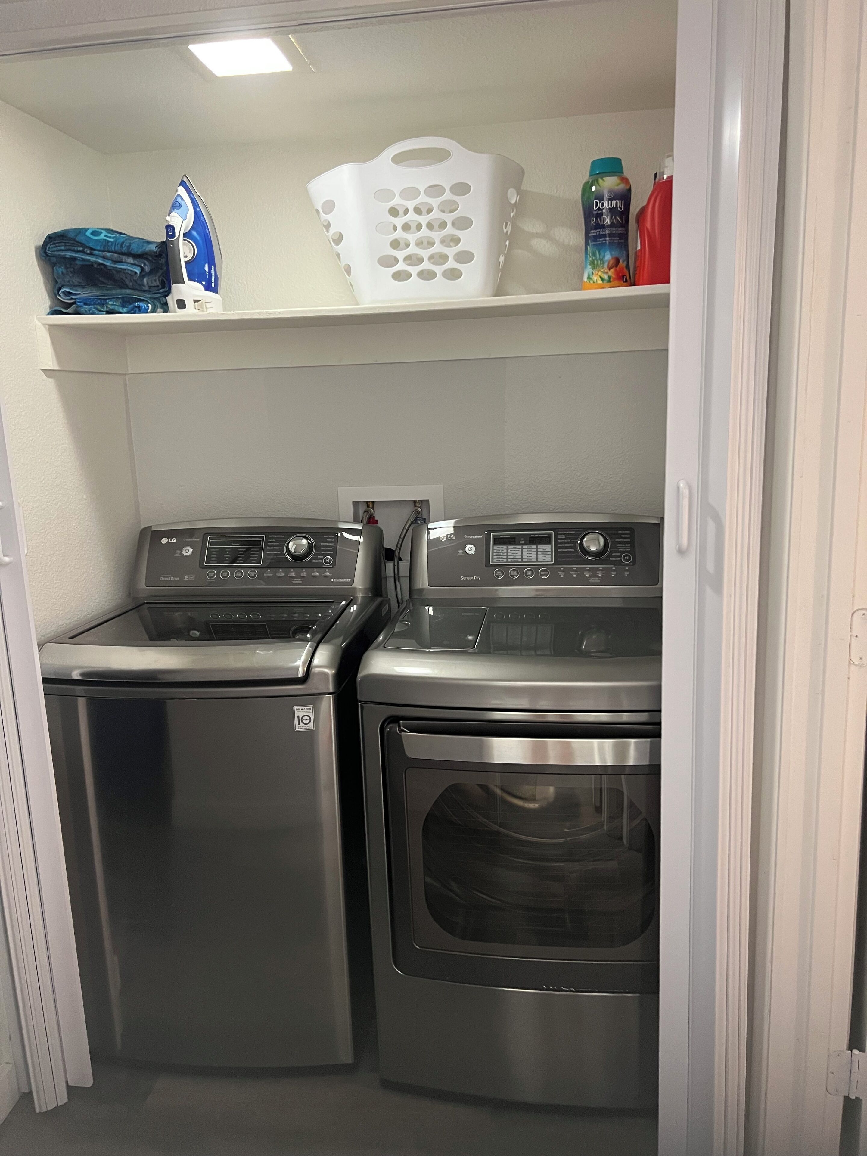 A laundry room with two stainless steel washers and dryers, a white basket, and a detergent bottle on a shelf.