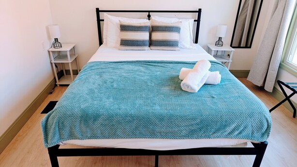 Un lit bien fait, recouvert d’une couverture turquoise, avec des oreillers blancs et des serviettes pliées. Il y a deux tables de chevet avec des lampes et une fenêtre avec des rideaux.