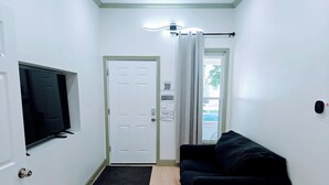 Une petite pièce propre, avec un canapé noir, un téléviseur fixé au mur, une porte avec une fenêtre et une petite fenêtre ornée d’un rideau.