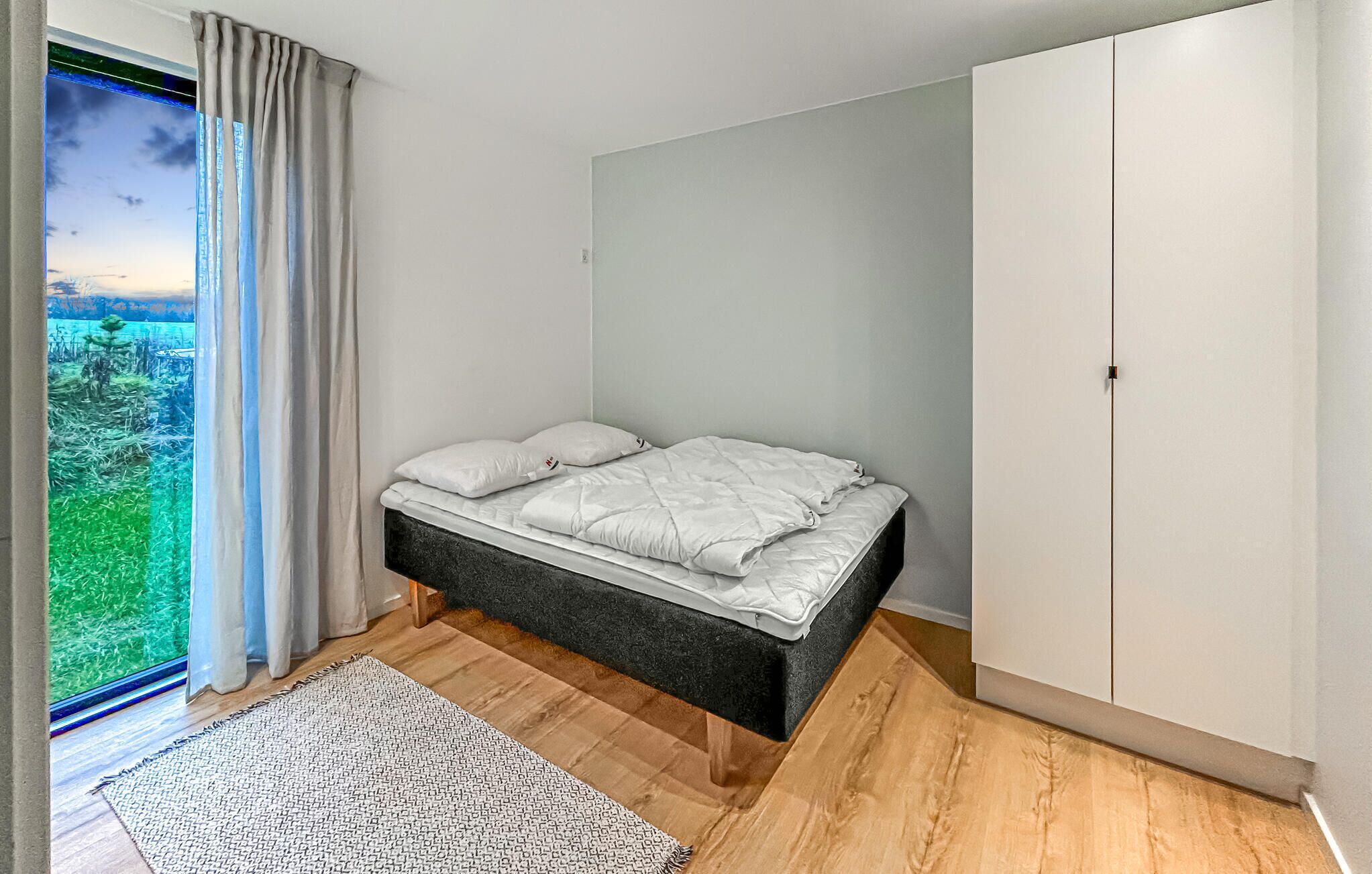 A bedroom with a bed, a wardrobe, a window with curtains, and a view of greenery outside.