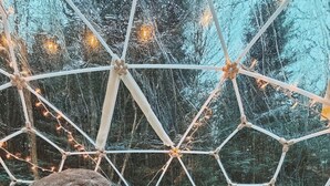 Un rincón acogedor con un sofá y cojines, una mesa con galletas y una taza, y vistas a través de una estructura en forma de cúpula geodésica con luces colgantes.