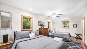 Un dormitorio con dos camas, un ventilador de techo, un aparador y una ventana con vistas a árboles.