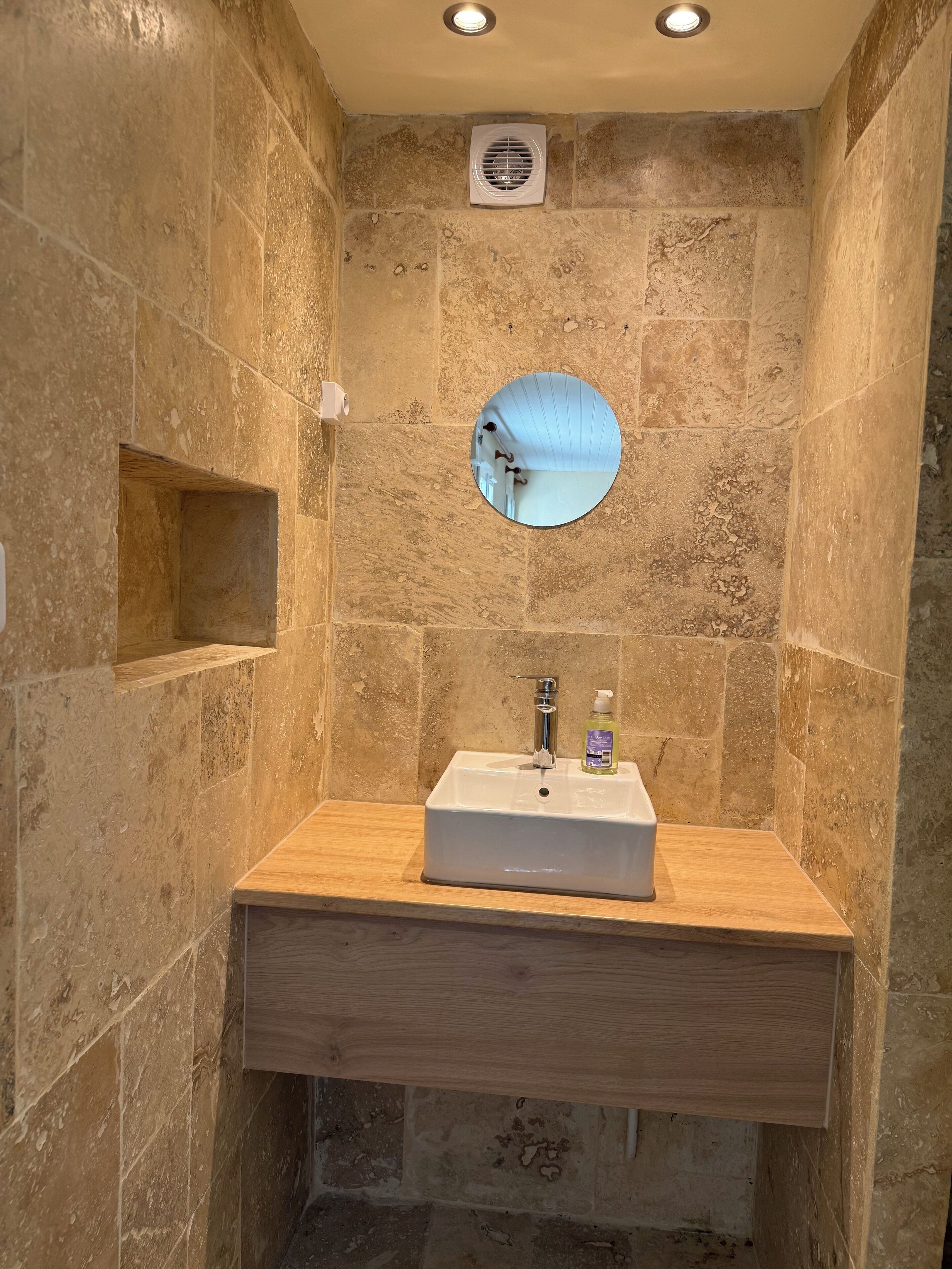 Un baño con pared de azulejos color crema, tocador de madera, lavabo blanco, espejo redondo e iluminación en el techo.