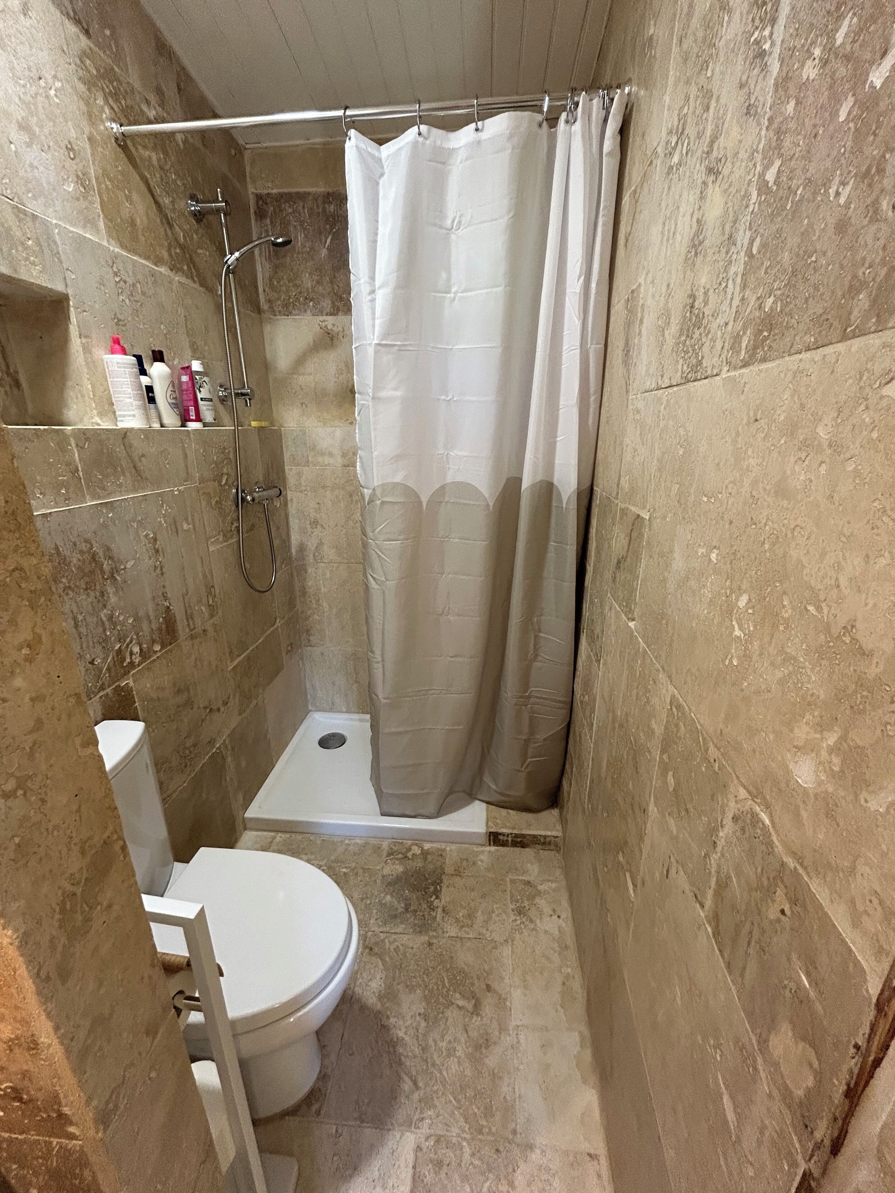 Une petite salle de bain carrelée, équipée d’une douche, de toilettes et d’une étagère où sont rangés les articles de toilette.
