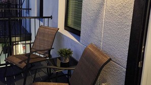 Un balcón con sillas de mimbre, una mesita con una planta, una ventana con persianas y un farol fijado a la pared.