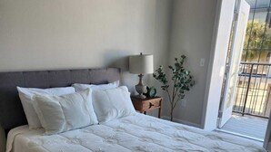 Una cama grande con cabecera gris, ropa de cama blanca y una mesita de noche con una lámpara y una planta.